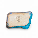 A handmade ceramic clay dish with a rustic turquoise blue glazed edge and a beige sandy center, resting on a dark wood surface against a light green wall. The center features a stamped logo reading 'Country Clay Clay'.