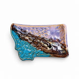 A handmade ceramic tray shaped like the state of Montana, featuring a glossy glaze with turquoise blue, purple, brown, and gold colors, displayed on a wooden surface.
