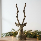Decorative deer head sculpture with antlers on a wooden surface with Christmas lights and greenery in the background.