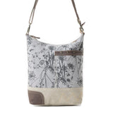 A slouchy shoulder bag with a gray and black botanical dandelion print on the upper half, a beige canvas lower half with a gold embroidered 'M', and brown leather trim and strap.