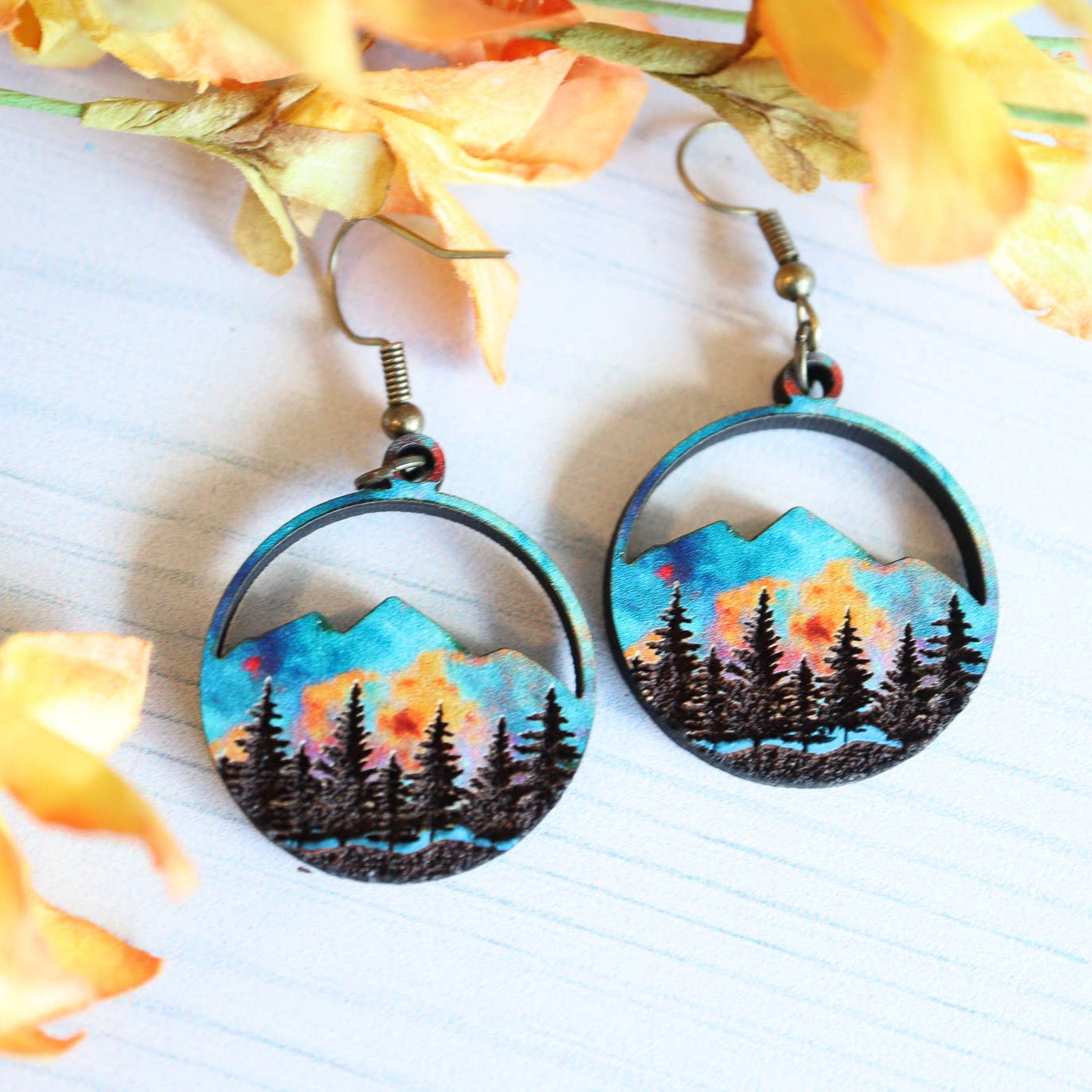 Pair of earrings with mountain and forest design on a white surface with yellow flowers.