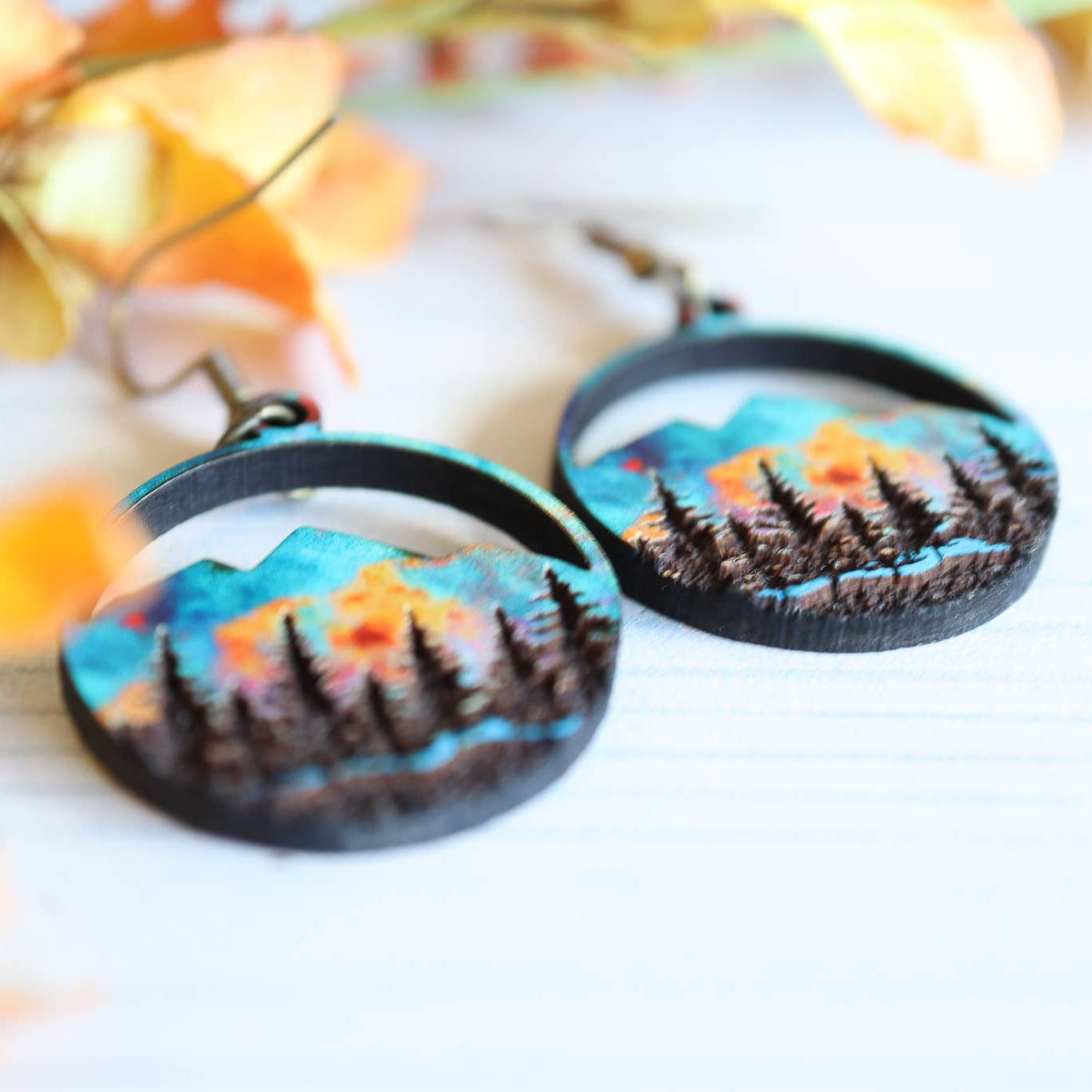 Pair of earrings with mountain and forest design on a white background with flowers.