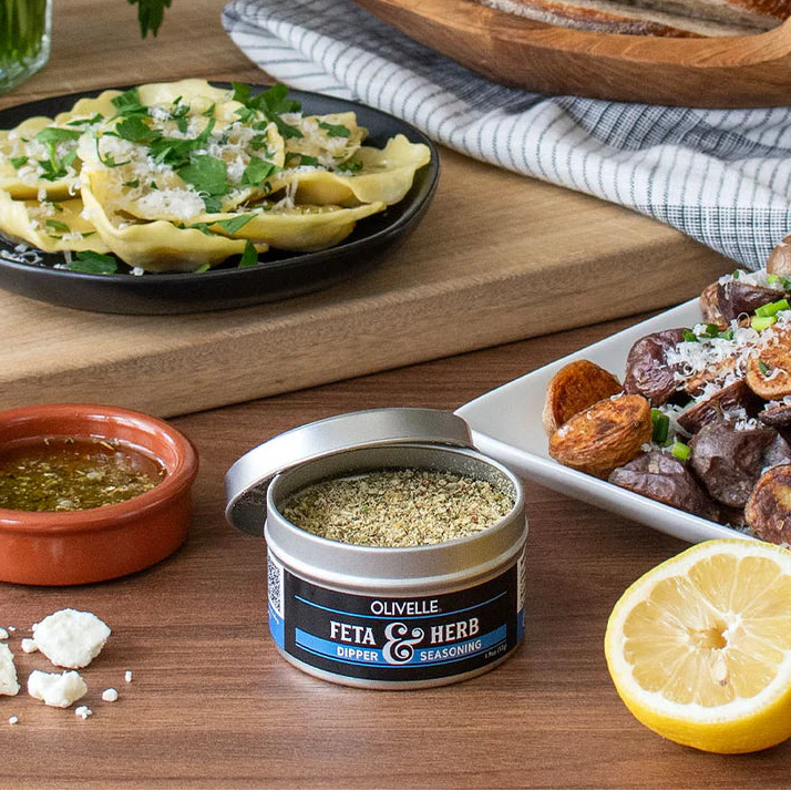 Table with food items including a bowl of pasta, bread, and a container of Olivelle Feta & Herb seasoning.