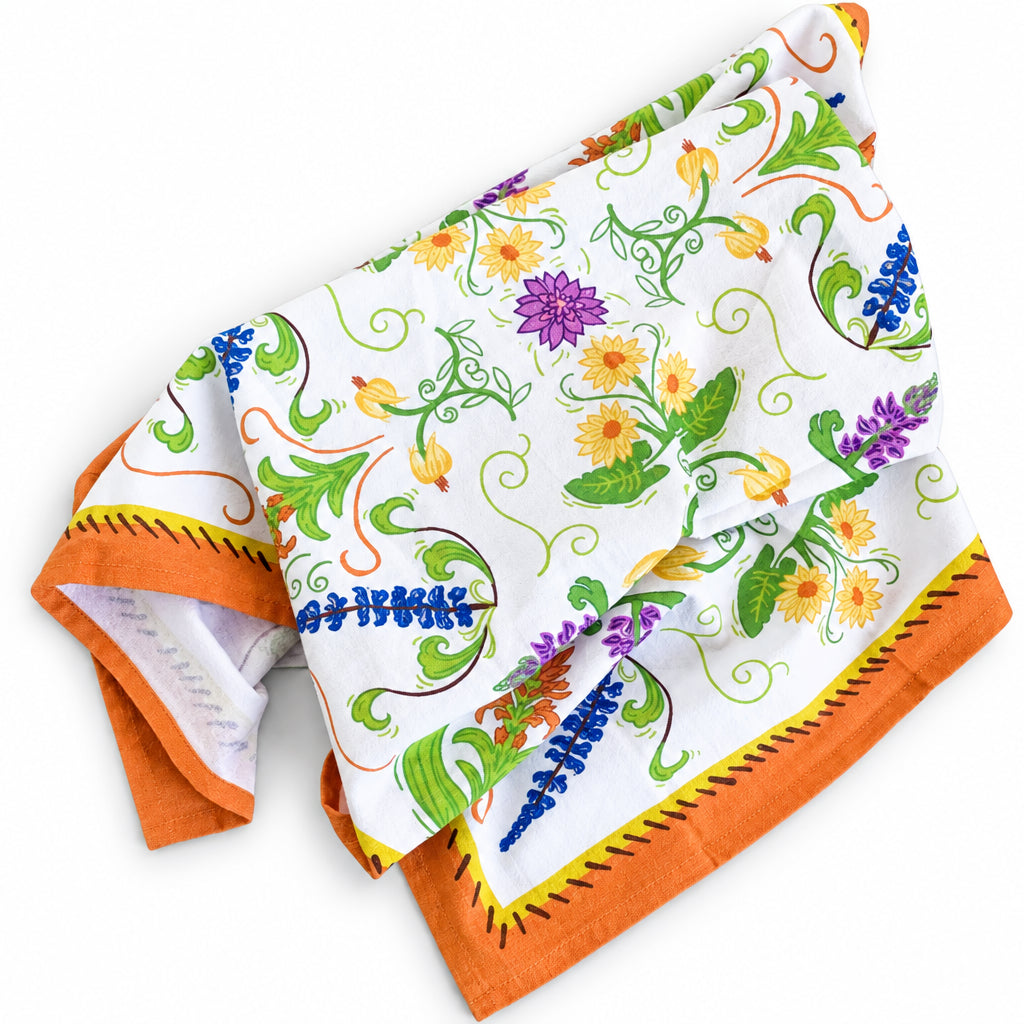 A square white cotton kitchen towel with a colorful floral and scroll pattern, finished with an orange border and yellow stitched trim, laid flat on a white surface.