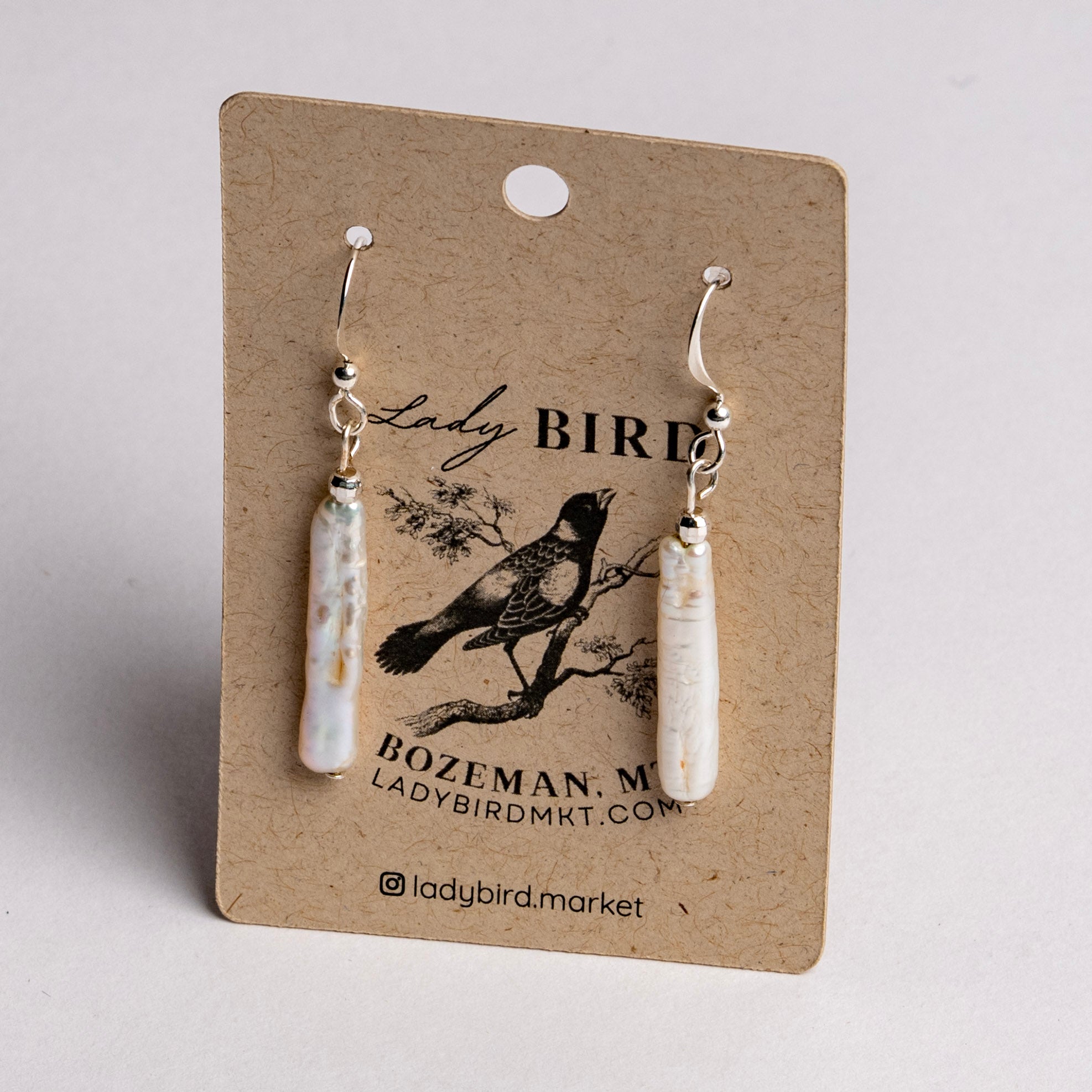 A pair of white pearl dangle earrings on a brown kraft display card with a black bird illustration and the text Lady Bird, Bozeman, MT, Ladybirdmk.com, photographed against a light gray background.