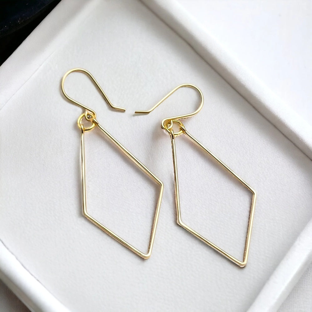 A pair of gold geometric drop earrings with open diamond-shaped frames and fishhook ear wires, displayed on a white textured surface.