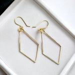 A pair of gold geometric drop earrings with open diamond-shaped frames and fishhook ear wires, displayed on a white textured surface.