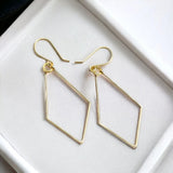A pair of gold geometric drop earrings with open diamond-shaped frames and fishhook ear wires, displayed on a white textured surface.