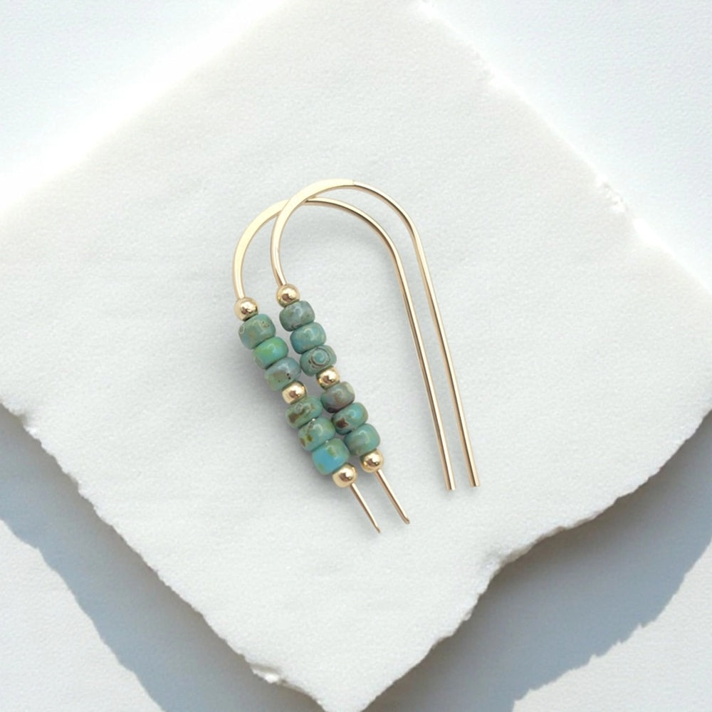 A pair of gold-tone double-ear wire earrings with small green marbled beads and gold spacer beads, displayed on a white stone slab.