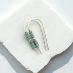 A pair of gold-tone double-ear wire earrings with small green marbled beads and gold spacer beads, displayed on a white stone slab.
