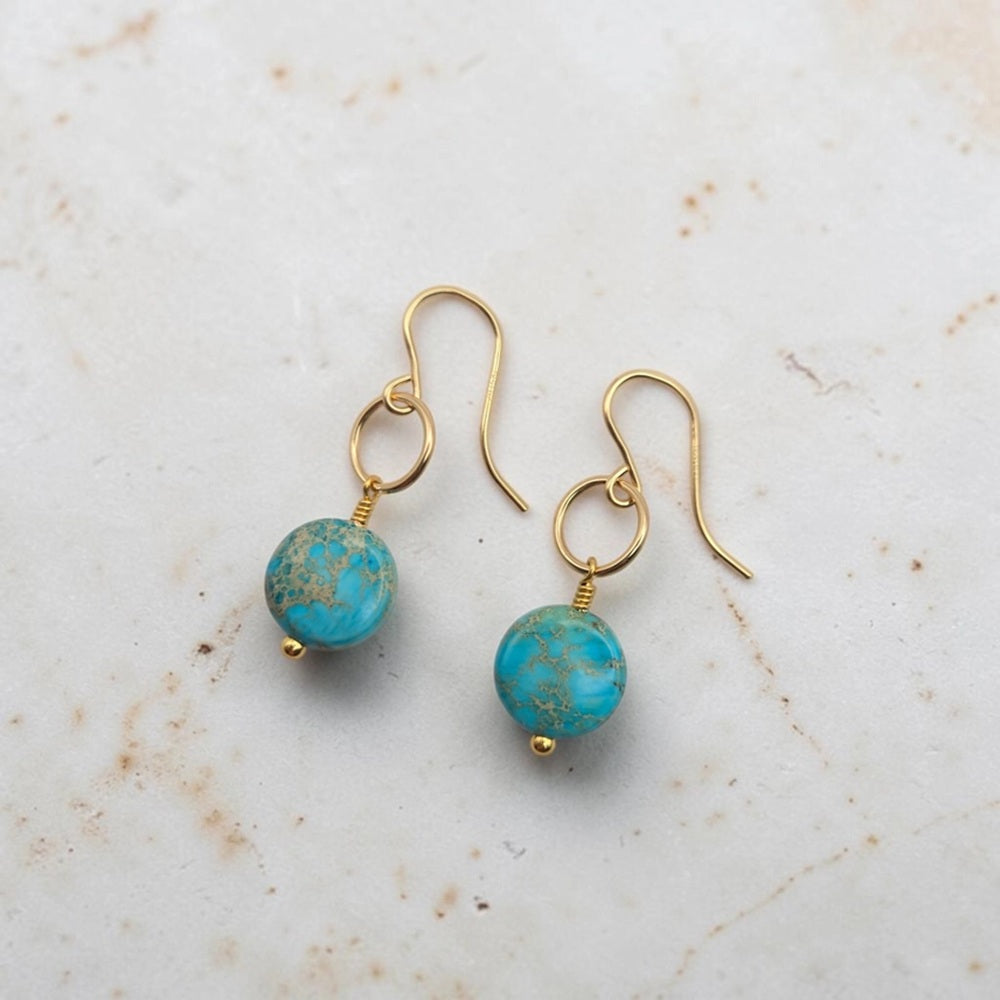 A pair of gold-tone dangle earrings with round turquoise-blue beads featuring gold flecks, displayed on a light beige marble surface.