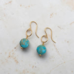 A pair of gold-tone dangle earrings with round turquoise-blue beads featuring gold flecks, displayed on a light beige marble surface.