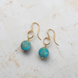 A pair of gold-tone dangle earrings with round turquoise-blue beads featuring gold flecks, displayed on a light beige marble surface.