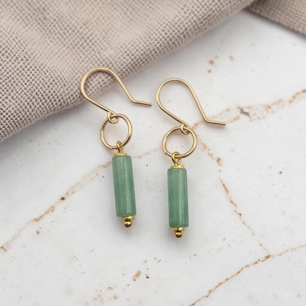 A pair of gold-tone dangle earrings with cylindrical light green beads, displayed on a white marble surface with a beige fabric napkin in the background.