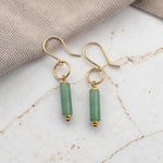 A pair of gold-tone dangle earrings with cylindrical light green beads, displayed on a white marble surface with a beige fabric napkin in the background.