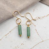 A pair of gold-tone dangle earrings with cylindrical light green beads, displayed on a white marble surface with a beige fabric napkin in the background.