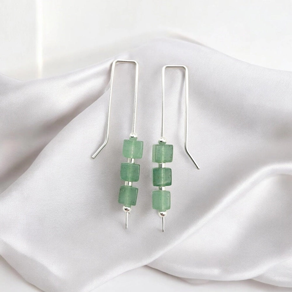 A pair of silver dangle earrings with three green faceted beads on each earring, displayed on draped white satin fabric.