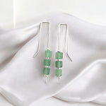 A pair of silver dangle earrings with three green faceted beads on each earring, displayed on draped white satin fabric.