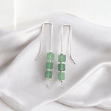 A pair of silver dangle earrings with three green faceted beads on each earring, displayed on draped white satin fabric.
