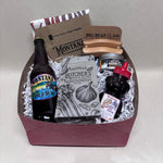 A curated gift basket in a maroon box filled with silver crinkle paper, containing a bottle of Montana Craft Ale BBQ sauce, a package of Olivelle's Butcher's Blend, a jar BBQ rub, a dish towel, a set of meat pullers, and a small bottle of honey with a red lid.