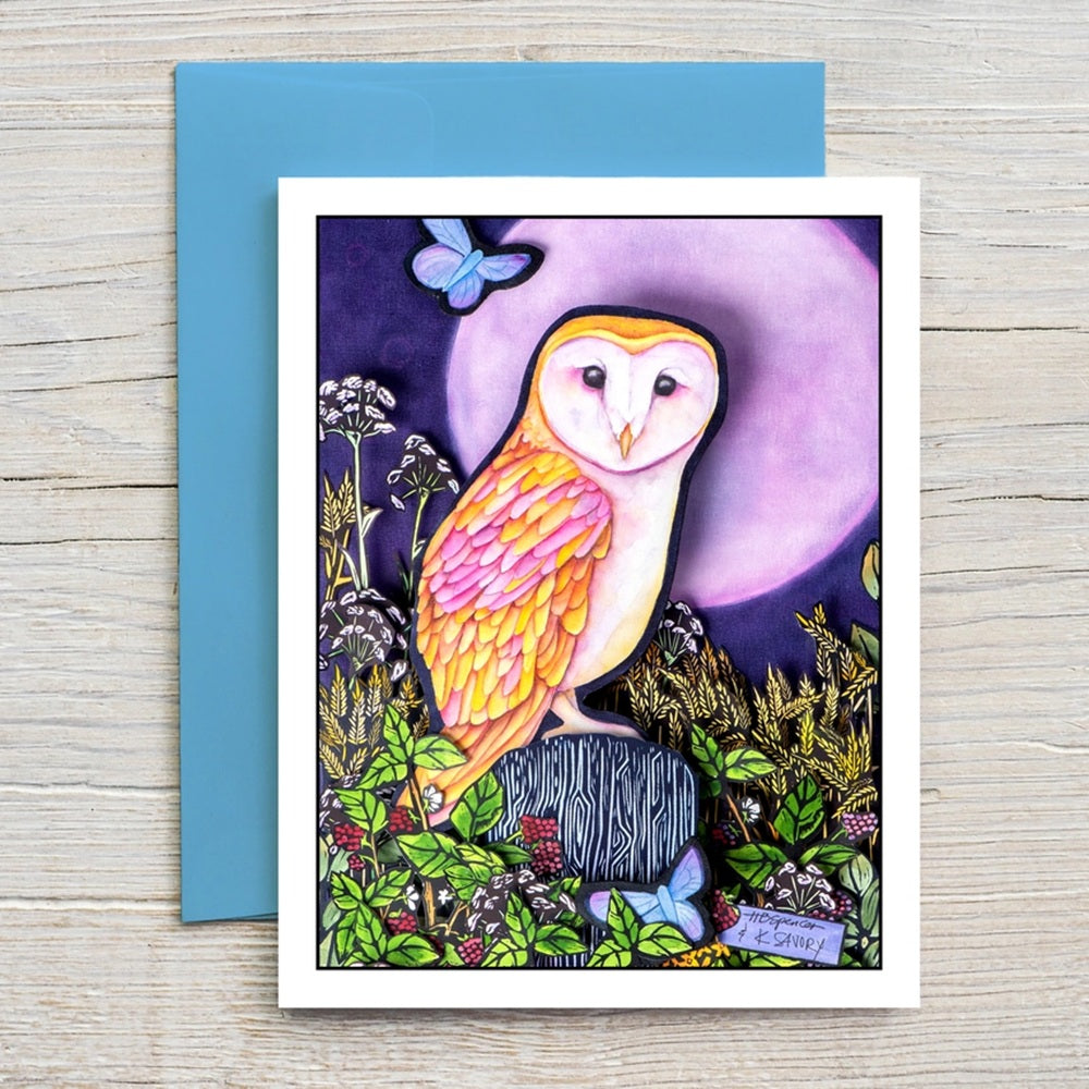 A square art print or greeting card featuring a colorful barn owl perched among flowers and foliage in front of a large purple moon, with a blue butterfly and a blue envelope behind it on a light wood surface.