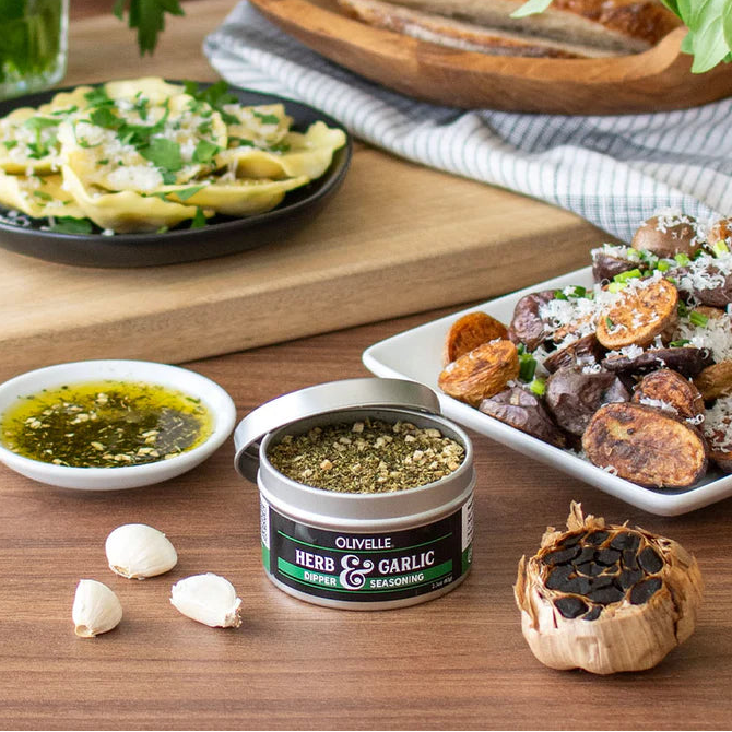 Table with food, herbs, and a container of Olivelle herb and garlic seasoning.