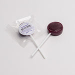 A single round dark berry lollipop with a white stick, shown next to its clear plastic wrapper and white circular label that reads 'Huckleberry'.