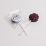 A single round dark berry lollipop with a white stick, shown next to its clear plastic wrapper and white circular label that reads 'Huckleberry'.