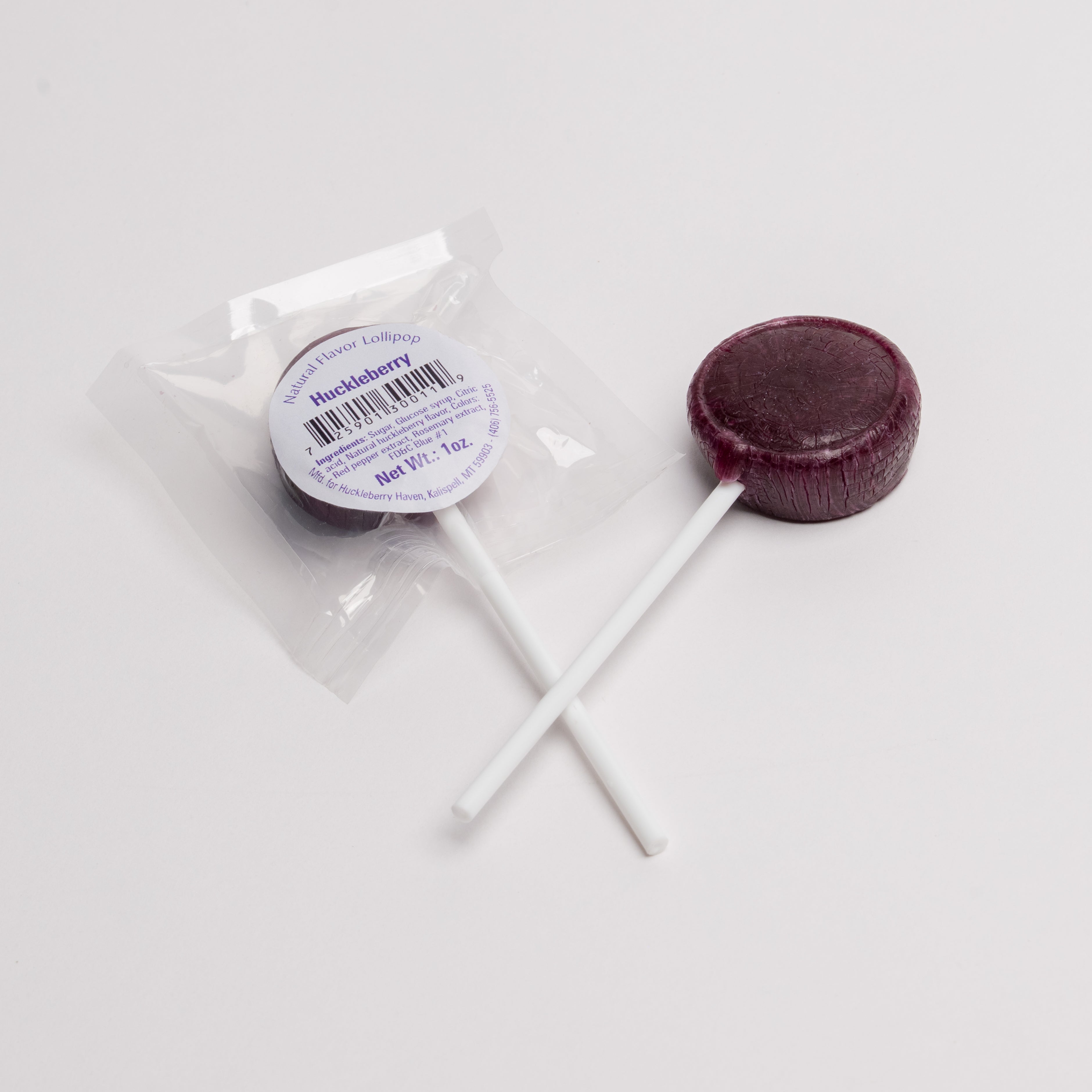 A single round dark berry lollipop with a white stick, shown next to its clear plastic wrapper and white circular label that reads 'Huckleberry'.