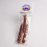 A packaged bag of rock candy on a plain light background. The clear plastic bag contains three long wooden sticks coated with dark purple crystalline candy, and the top card reads 'Old Fashioned Wild Huckleberry Rock Candy' with purple mountain graphics.