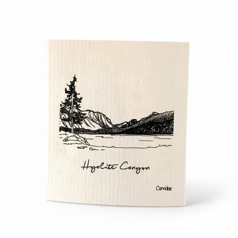 Dishcloth with a black and white illustration of a mountain landscape and text on a wooden surface.