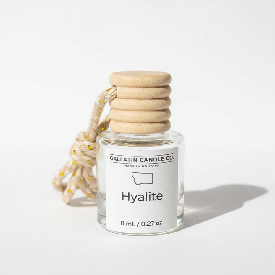 A small glass bottle candle with a white label reading 'GALLATIN CANDLE CO' and 'Hyalite', topped with a stacked beige wooden stopper and a braided beige cord with small gold beads, photographed on a light gray background.