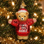 A teddy bear Christmas ornament hanging on a decorated Christmas tree. The ornament is made of textured red glass with gold trim, a red Santa hat with a white band, and pink glittery gloves.