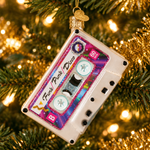 A cassette tape Christmas ornament hanging from a gold loop on a decorated Christmas tree with warm golden lights and tinsel in the background.