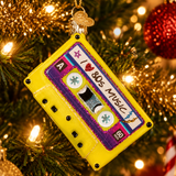 A yellow cassette tape Christmas ornament hanging on a lit Christmas tree. The ornament features purple glitter borders, retro label graphics, and a gold hanging loop with a small gold ornament.