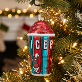 A Christmas ornament shaped like a retro iced coffee cup hanging on a decorated Christmas tree with warm string lights.