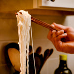 Person holding a strand of pasta with chopsticks in a kitchen setting