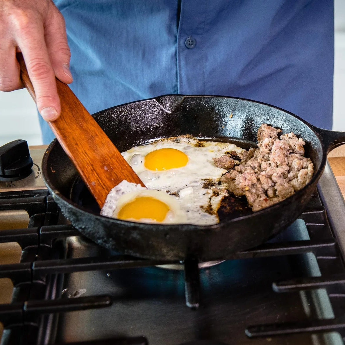 Person cooking eggs and sausage in a frying pan on a stove with eggs on a plate nearby.