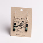 A pair of dangling earrings displayed on a kraft paper card. The earrings have silver fishhook wires and cylindrical beads featuring black, turquoise blue, and white stripes, with a small black bird illustration in the center.