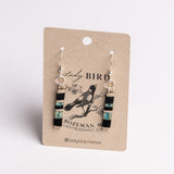 A pair of dangling earrings displayed on a kraft paper card. The earrings have silver fishhook wires and cylindrical beads featuring black, turquoise blue, and white stripes, with a small black bird illustration in the center.