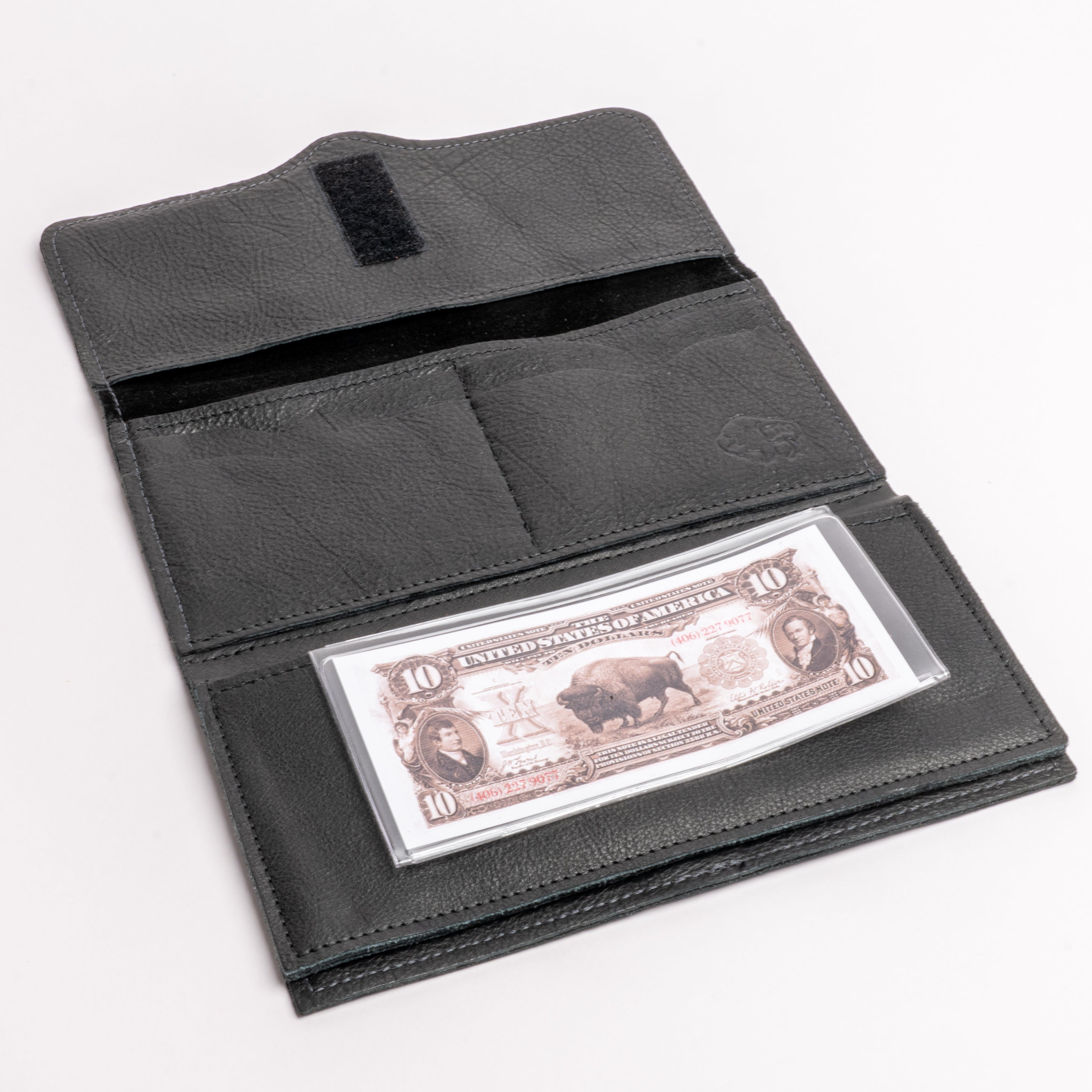 A black leather wallet shown open on a white background, displaying its interior compartments, a black hook-and-loop closure tab, and a clear plastic ID window holding a vintage-style $10 bill.