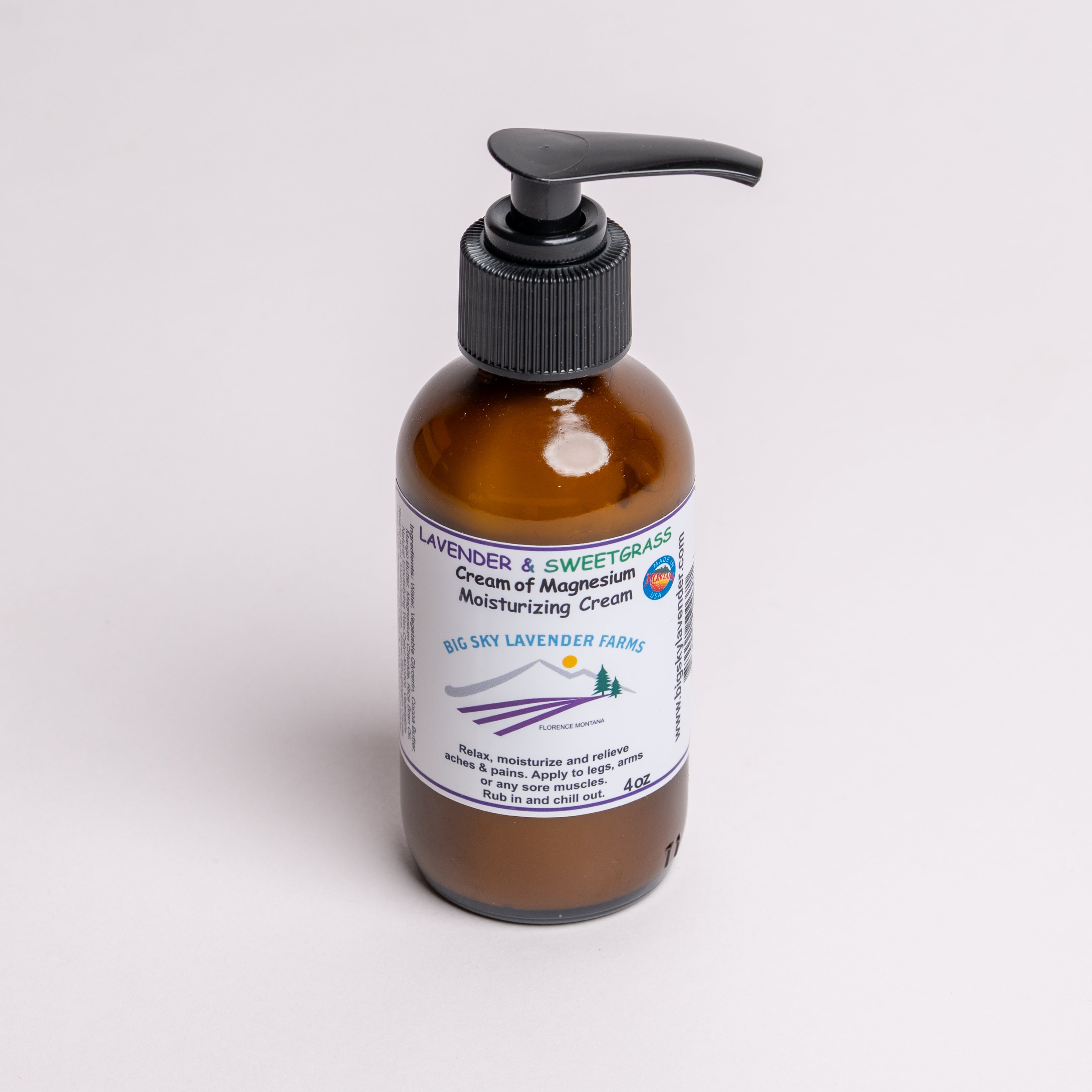 An amber glass pump bottle of Big Sky Lavender Farms Lavender & Sweetgrass Cream of Magnesium Moisturizing Cream, featuring a black pump dispenser and a white label with purple and blue text, set against a plain light gray background.