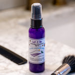 Purple spray bottle labeled 'Let it Be' on a bathroom counter with a phone and brush.