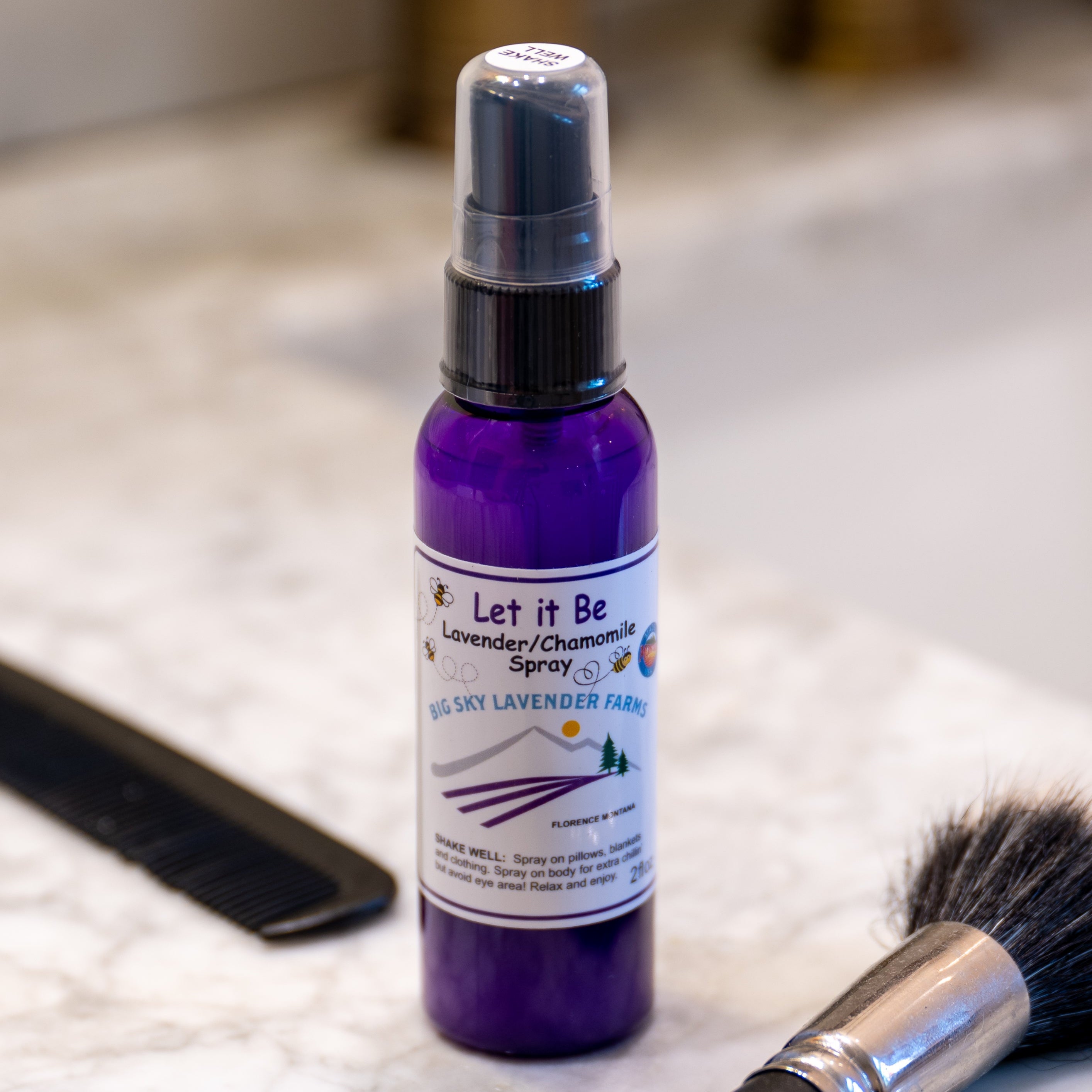 Purple spray bottle labeled 'Let it Be' on a bathroom counter with a phone and brush.
