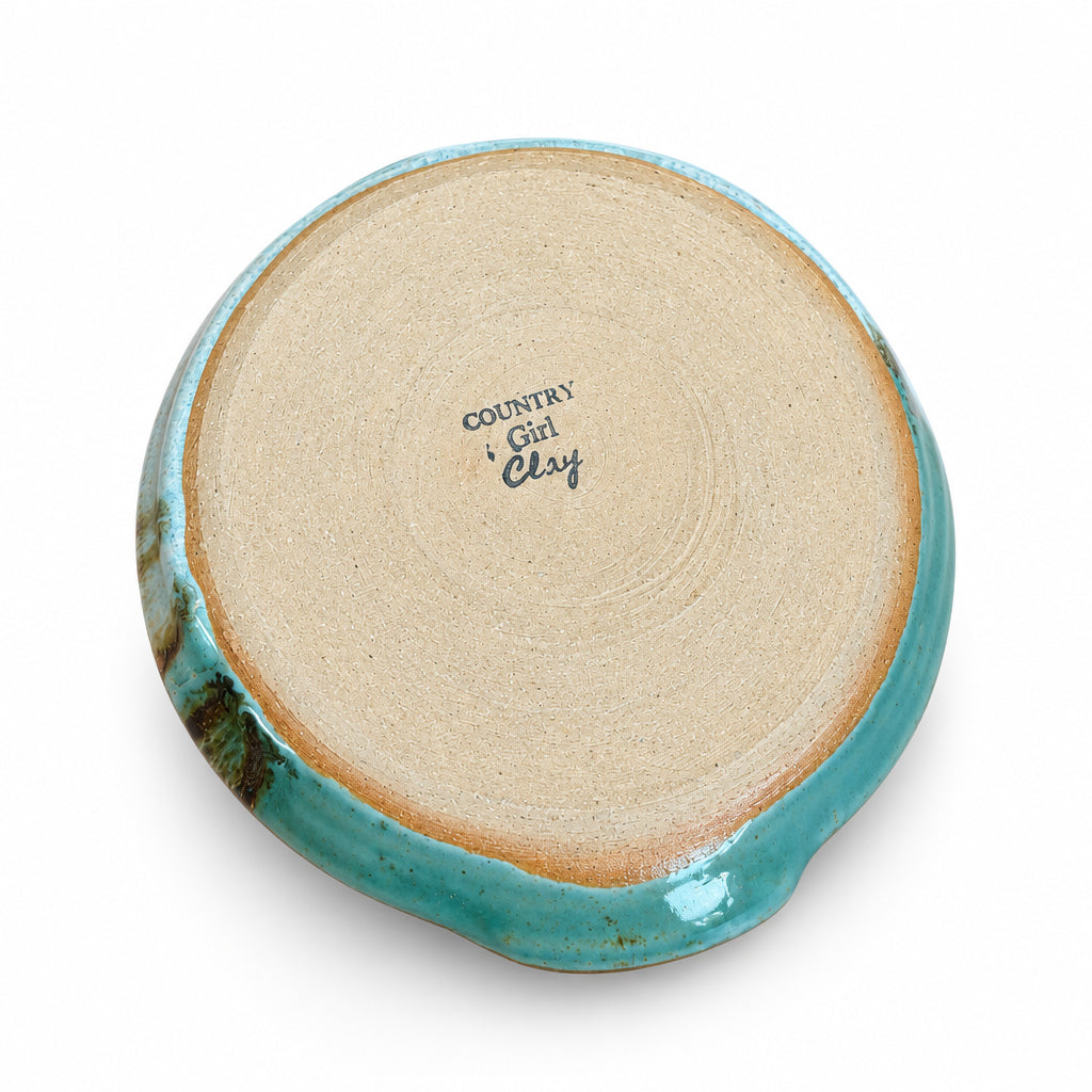 The bottom of a handmade ceramic plate or dish, featuring a light beige clay base with a glossy turquoise blue glaze along the rim and a small dark green patch on the left side. The brand name 'COUNTRY Girl Clay' is printed in black on the center.
