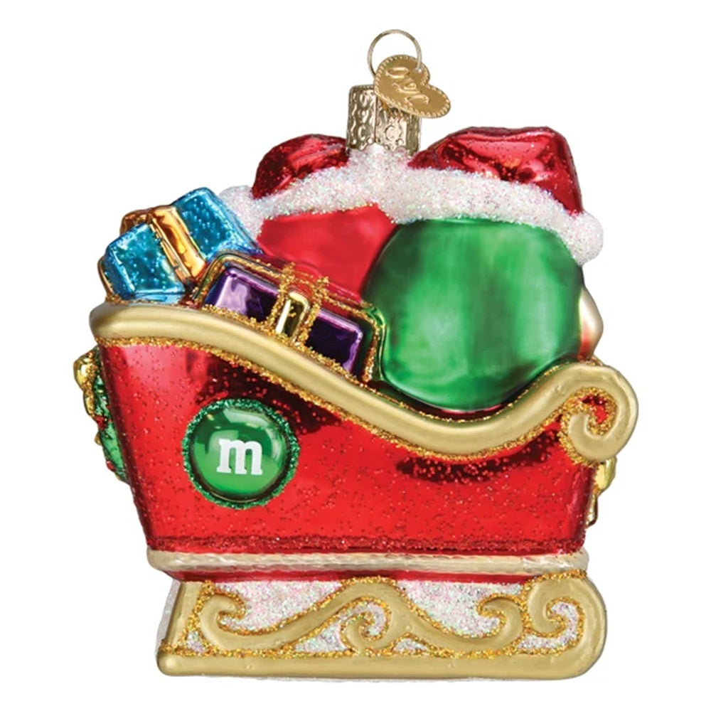 A Christmas ornament shaped like a red sleigh filled with gifts and Santa hats, with a green circle on the side featuring a white lowercase 'm' and a gold hanging loop at the top, shown on a plain white background.