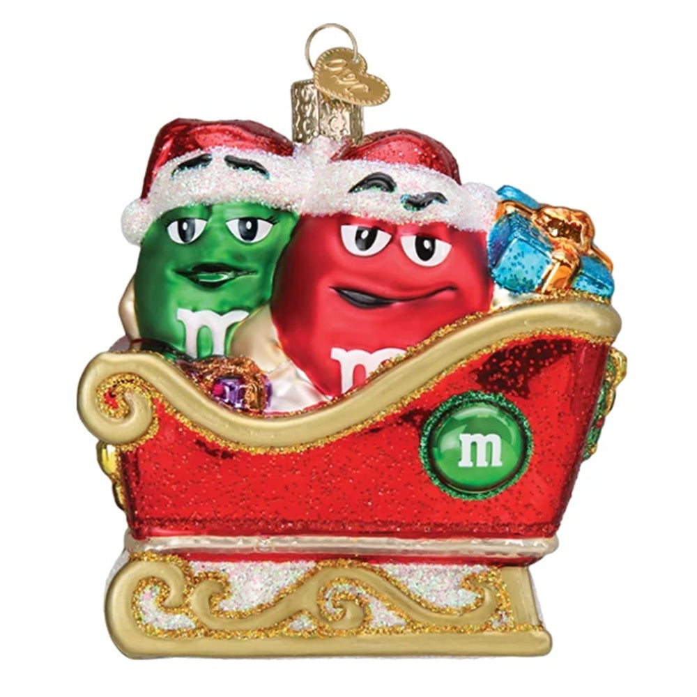 A Christmas ornament featuring two M&M's characters in a red sleigh. The green M&M is on the left wearing a Santa hat, and the red M&M is on the right with a Santa hat.