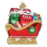 A Christmas ornament featuring two M&M's characters in a red sleigh. The green M&M is on the left wearing a Santa hat, and the red M&M is on the right with a Santa hat.