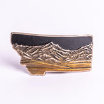 Metallic bronze belt buckle shaped like Montana with mountain design on a white background