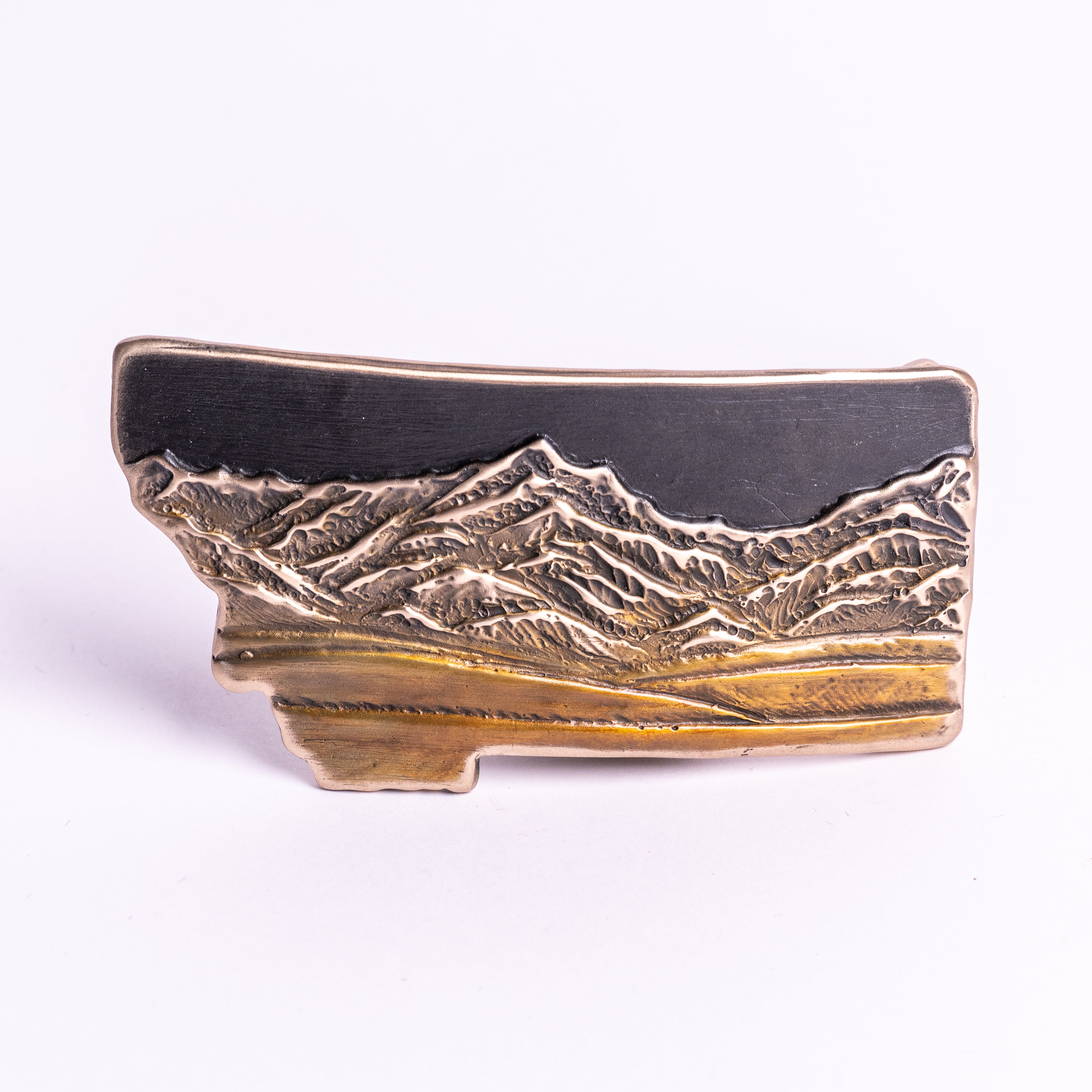 Metallic bronze belt buckle shaped like Montana with mountain design on a white background